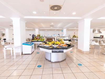 Salle de petit-déjeuner lumineuse avec buffet sur table centrale et chaises blanches.