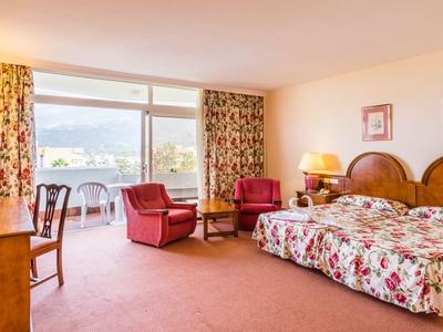 Chambre d'hôtel lumineuse avec grand lit, deux fauteuils et grande fenêtre avec vue sur balcon.