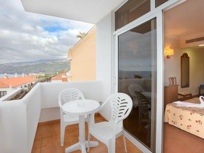 Balcon avec chaises blanches et table, vue sur montagnes et toits sous ciel nuageux.