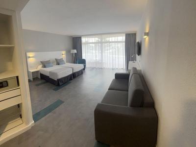 Chambre d'hôtel moderne avec lit double, canapé gris et grande fenêtre avec rideaux.