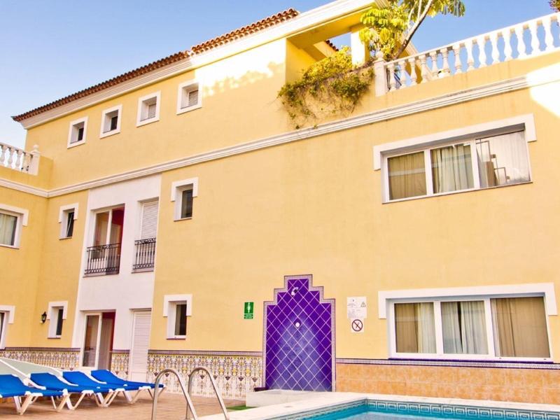 Žlutý hotel s modrým bazénem, lehátky a houpacími sítěmi pod jasnou oblohou.