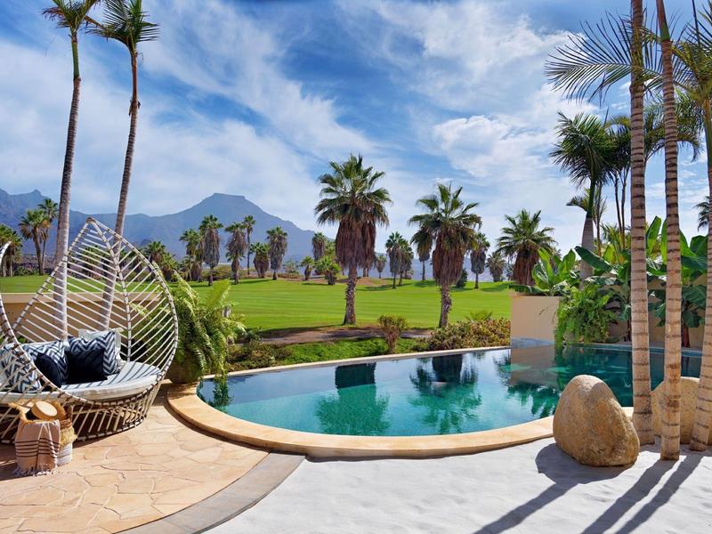 Luxusní bazén s palmami a zeleným trávníkem pod modrou oblohou.