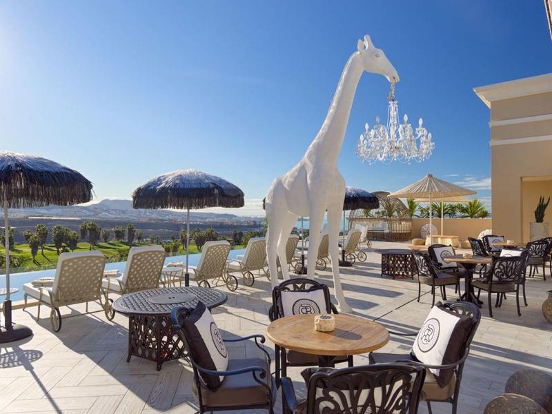 Bazénová terasa se sezením, slunečníky a bílou sochou žirafy s lustr.