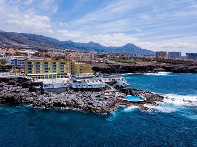 Hotel na skalistej linii brzegowej z basenem i górami w tle.