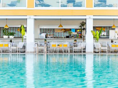 Piscine d'hôtel moderne avec un bar et des chaises en arrière-plan en plein jour.