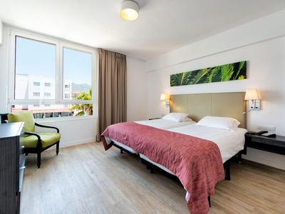 Chambre d'hôtel lumineuse avec lit double, fauteuil vert, grande fenêtre et sol en bois.