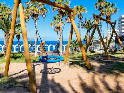 Plac zabaw z huśtawkami i palmami wzdłuż nadmorskiego bulwaru z widokiem na morze.