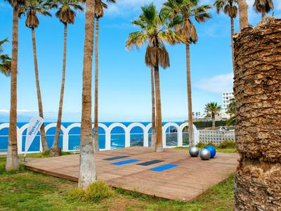 Espace de yoga en plein air avec plateforme en bois et vue sur la mer, entouré de palmiers et d'une rambarde blanche.