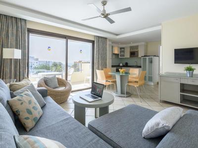 Helle Hotelzimmer-Lounge mit Sofa, Sesseln, Tisch und Küchenzeile mit Esstisch.
