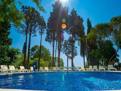 Runder Pool mit blauer Wasseroberfläche, umgeben von Liegestühlen und hohen Bäumen im Sonnenschein.