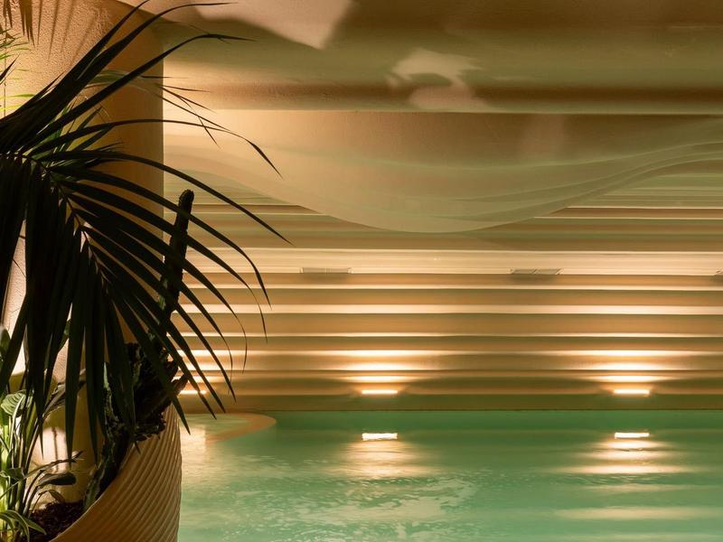 Innenpool mit warmem Wasser, beleuchtet von Wandleuchten, daneben ein Liegestuhl und Palmenblatt.