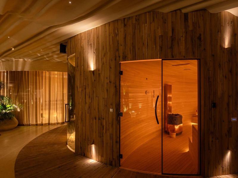 Innenraum mit warmem Licht, Holzwand, gebogenem Boden und offener Saunatür aus Holz und Glas.