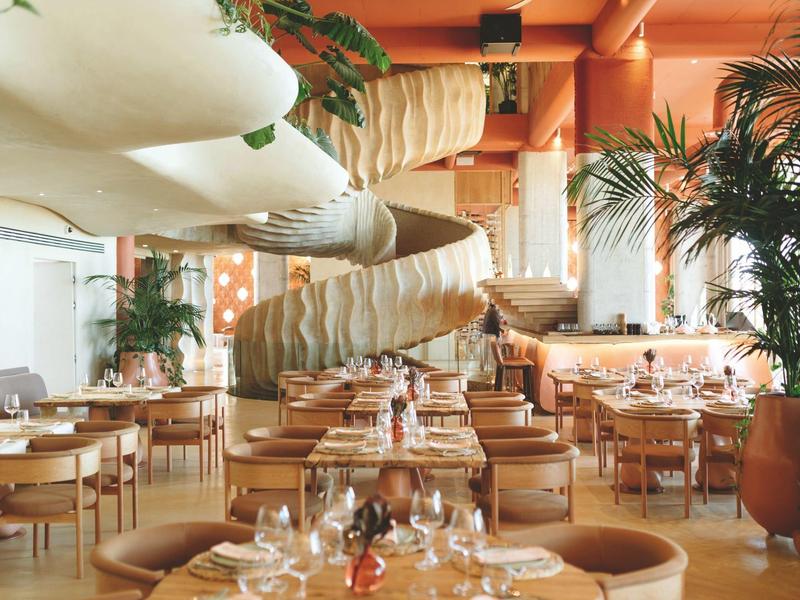 Hell eingerichtetes Restaurant mit Holztischen, Stühlen und vielen Pflanzen, helle Wände mit oranger Farbe.