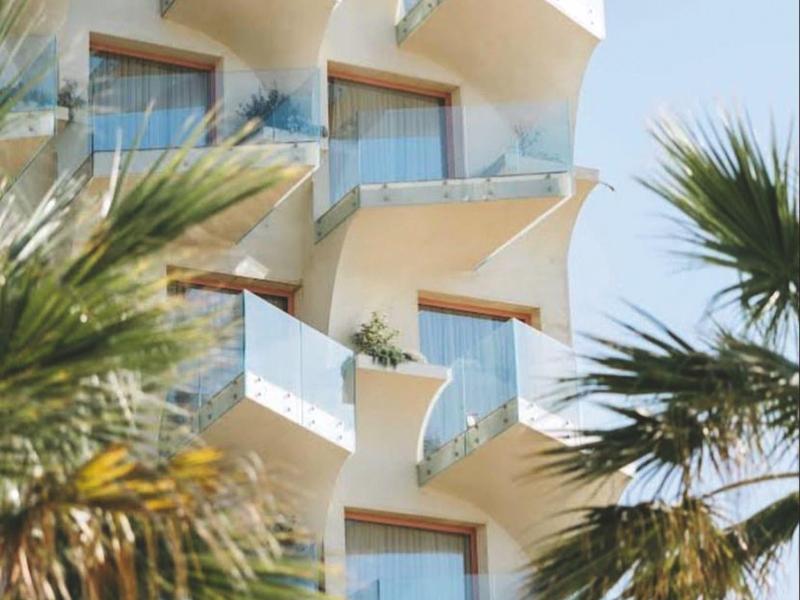 Moderner Wohnblock mit auffälligen, weißen Balkonen und Glasgeländern, von Palmen umgeben.