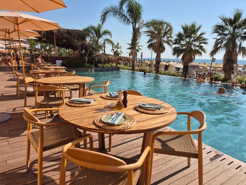 Poolterrasse mit runden Holztischen, Stühlen, Sonnenschirmen und Palmen im Hintergrund am Meer.