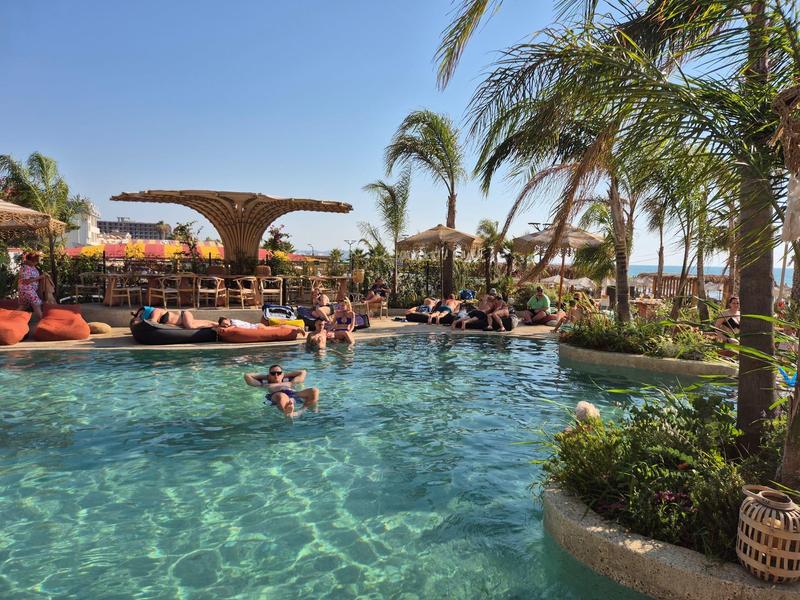 Pool mit klarem Wasser, Palmen und Sitzbereichen, Menschen entspannen, blauer Himmel.