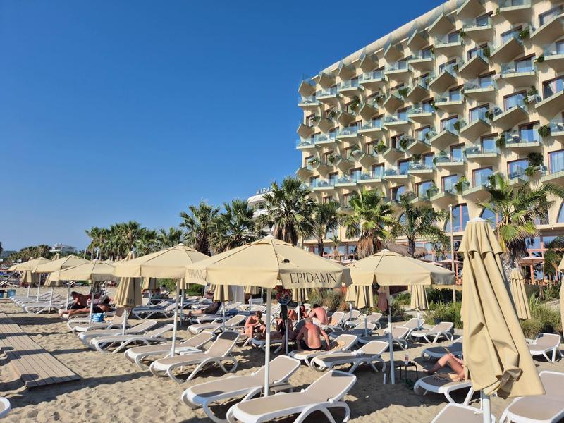 Strand mit Liegen, weißen Sonnenschirmen, Palmen und blauem Himmel neben einem Hotelgebäude.