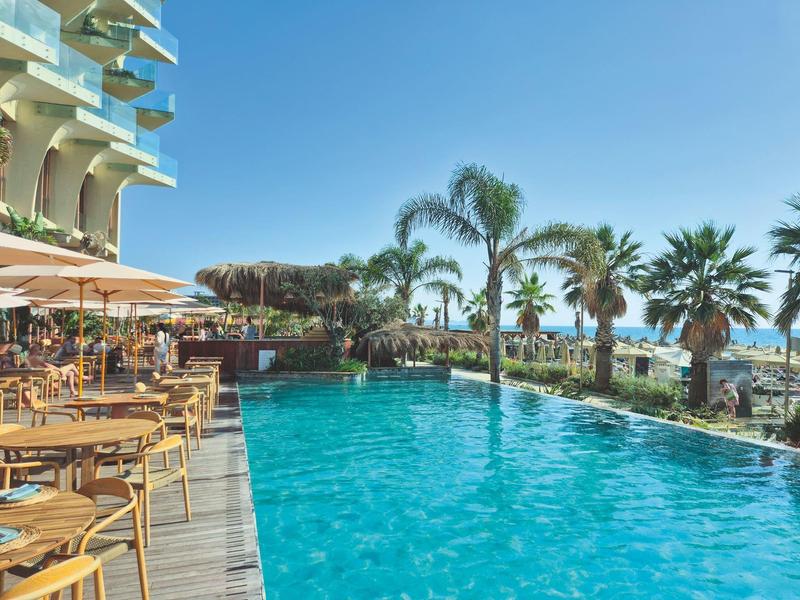 Schwimmbad mit klarem Wasser, umgeben von Palmen, Holzterrasse mit Tischen und Stühlen, sonniger Himmel.