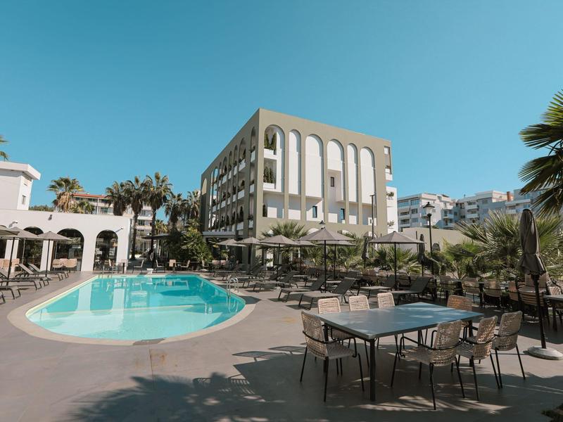 Moderner Hotelkomplex mit Pool, Außenbestuhlung, Palmen und klarem Himmel im Hintergrund.