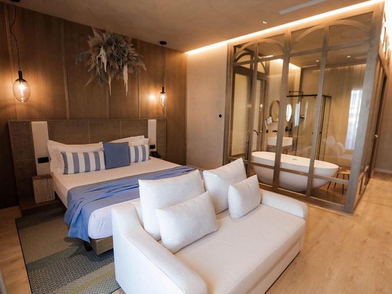 Modernes Hotelzimmer mit Doppelbett, weißem Sofa, Glasdusche und warmen Holzwänden.