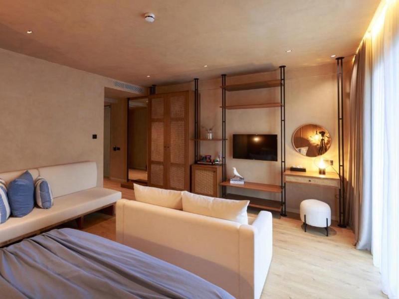 Modernes Hotelzimmer mit großem Bett, Sofa, TV an Wandregal und Tageslicht durch Vorhänge.