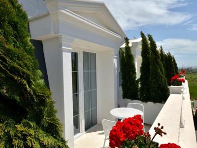 Balkon s bílými zdmi, červenými květinami a zelenými keři pod modrou oblohou.