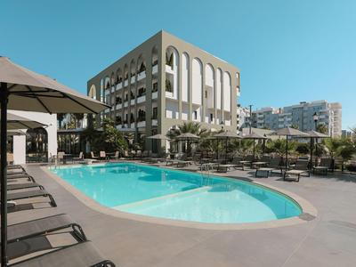 Moderní venkovní bazén s lehátky a slunečníky před výraznou budovou hotelu.