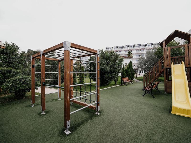 Kinderspielplatz mit Klettergerüst, Rutsche und grüner Rasenfläche, umgeben von Bäumen.