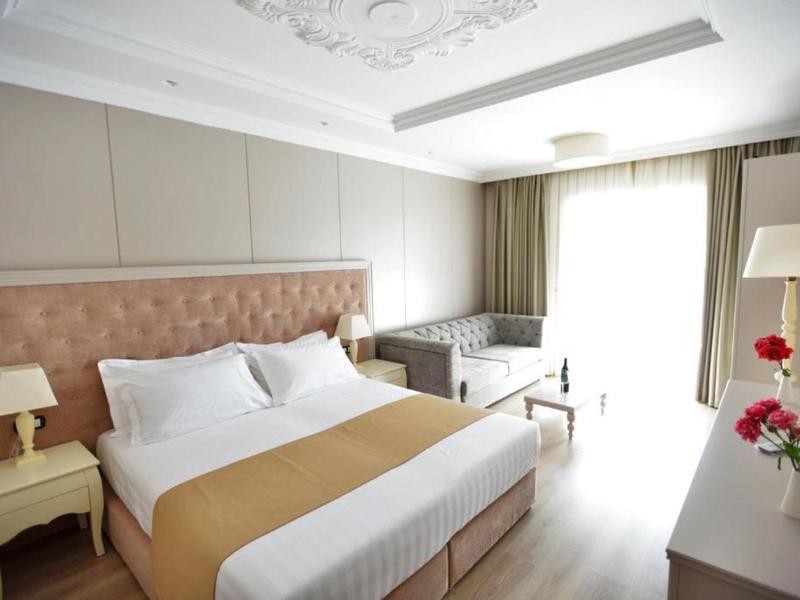 Helles Schlafzimmer mit Doppelbett, beiger Tagesdecke, Nachttischen, Sesseln und großen Fenstern mit Gardinen.