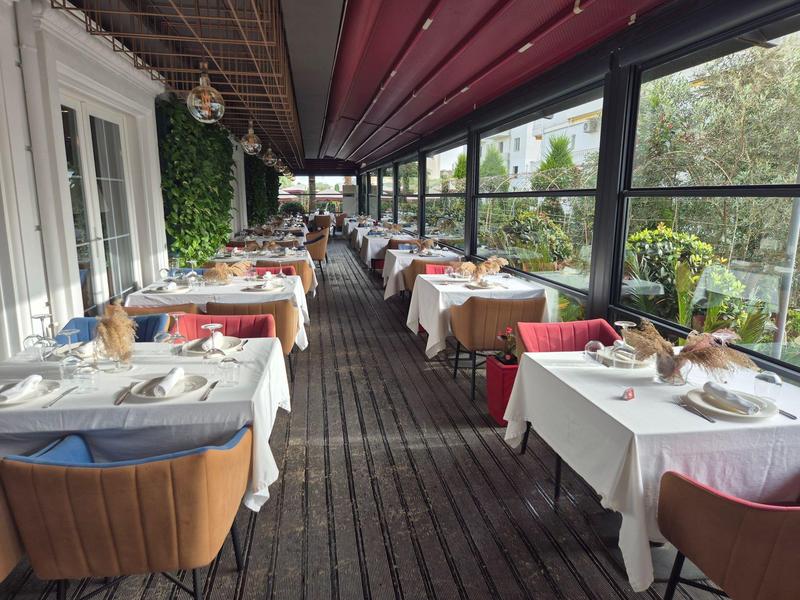 Überdachte Außenterrasse mit Holzboden, gedeckten Tischen und roten sowie braunen Sesseln.