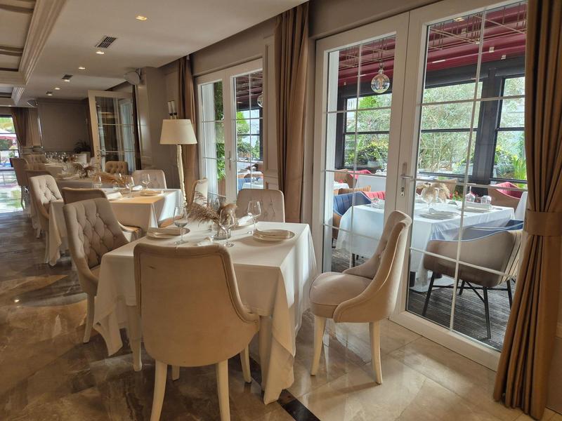 Elegantes Restaurant mit weißen Tischtüchern, bequemen Stühlen und großen Fenstern zum Garten hin.