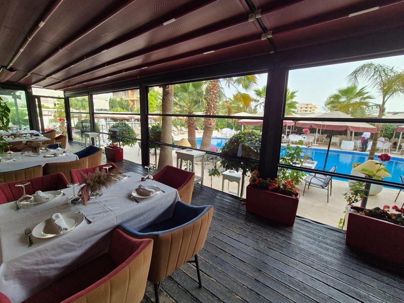 Überdachte Terrasse mit Holzboden, gedeckten Tischen und Blick auf Pool und Palmen im Hintergrund.