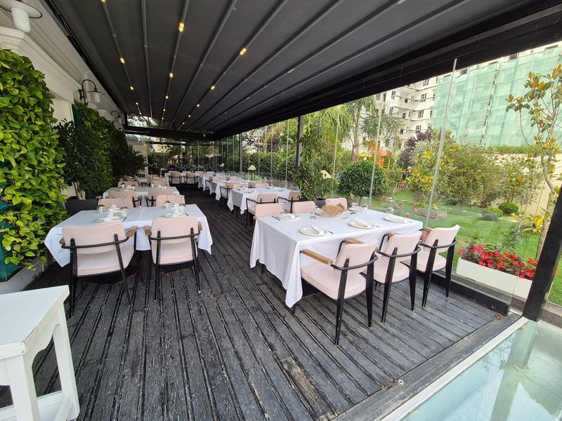 Überdachte Terrasse mit Holzboden, weißen Tischtüchern und Stühlen neben grüner Gartenanlage.