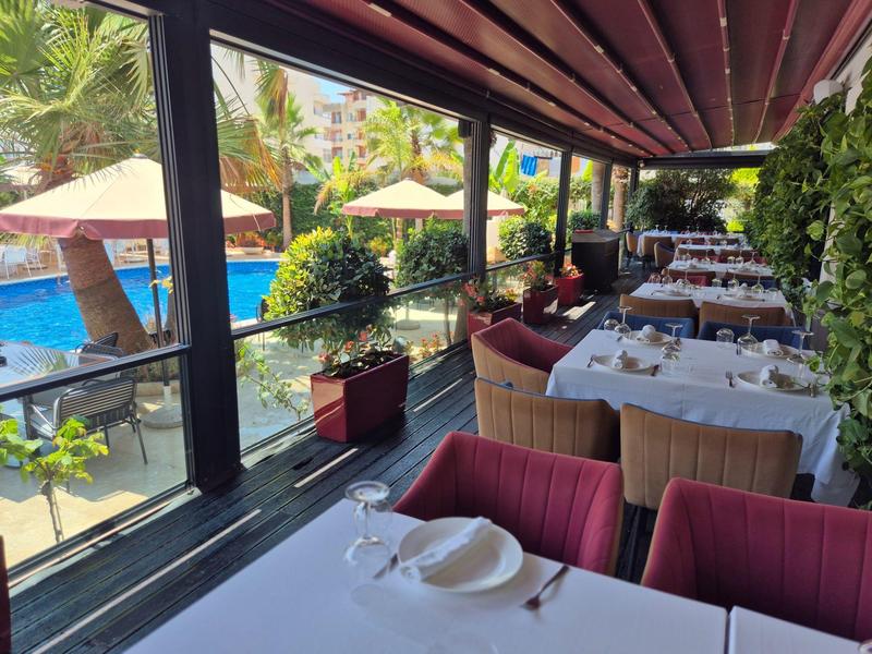 Terrassenrestaurant mit weißen Tischtüchern, roten Sesseln und Blick auf blauen Pool und Palmen.