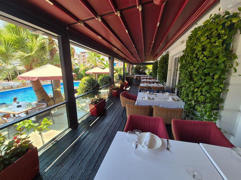 Terrassenrestaurant mit weißen Tischen, roten Stühlen, Pflanzen und Blick auf einen Pool mit Sonnenschirmen.