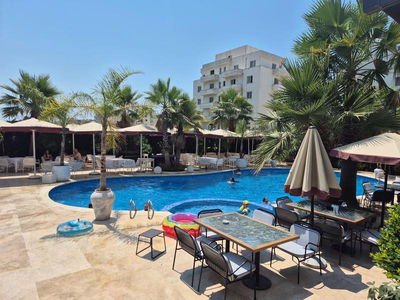 Außenpool mit Liegestühlen, Tischen, Sonnenschirmen und Palmen vor einem mehrstöckigen Gebäude.