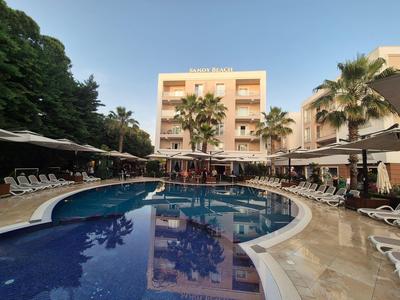 Hotel mit Außenpool und Sonnenliegen, umgeben von Palmen und blauem Himmel.