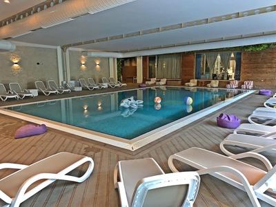 Innenraum mit einem rechteckigen Pool, umgeben von weißen Liegestühlen und Holzboden.