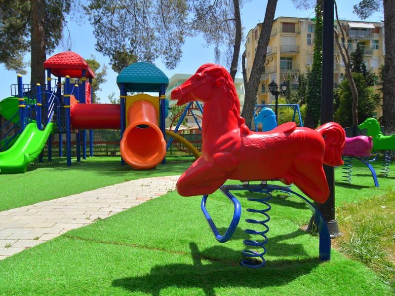 Bunter Spielplatz mit Rutschbahnen und einem roten Federpferd im Gras nahe einem Hotel.