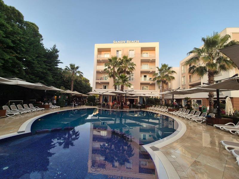 Hotel mit Außenpool und Sonnenliegen, umgeben von Palmen und blauem Himmel.