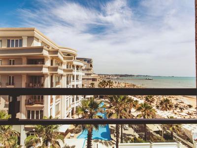 Blick von Balkon auf Hotel, Palmen, Pool und Strand mit Meer unter blauem Himmel.