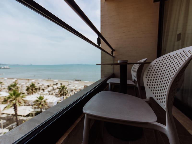 Balkon se židlí a stolem s výhledem na pláž a moře pod jasnou oblohou.
