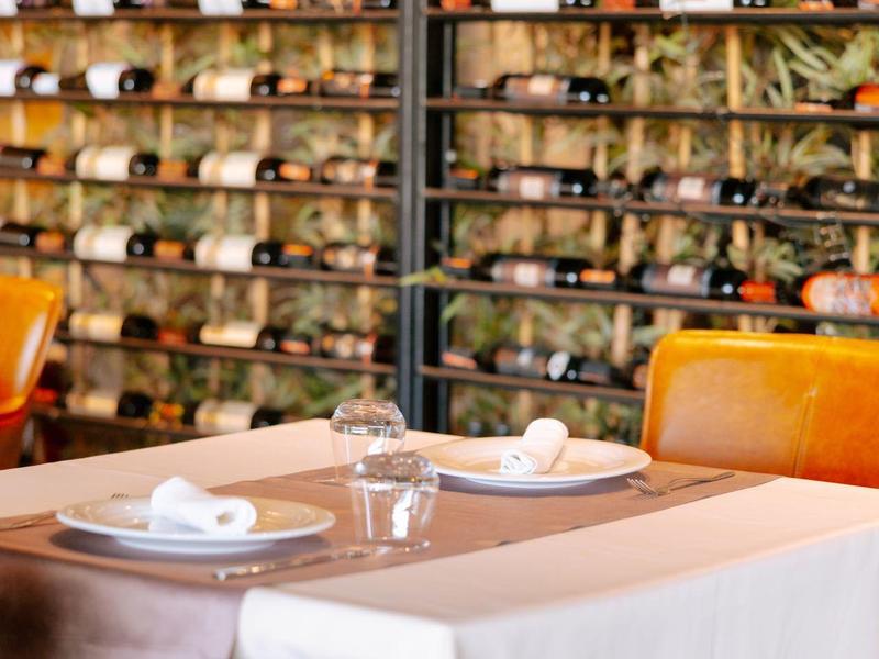 Elegantní jídelní stůl s bílými ubrousky a vinnými sklenicemi před zdí s lahvemi vína.