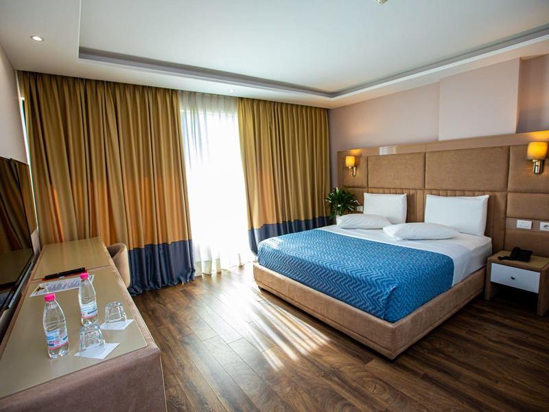 Moderní hotelový pokoj s manželskou postelí, dřevěnou podlahou a velkými okny s závěsy.