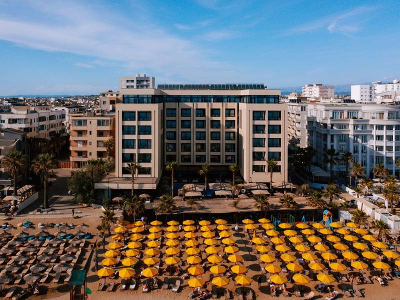 Pohled na hotel s pláží plnou žlutých slunečníků a lehátek.