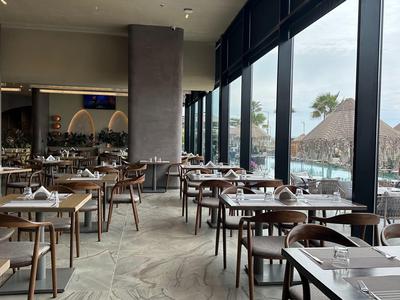 Modernes Restaurant mit Holzstühlen und großen Fenstern mit Blick auf Palmen und Sonnenschirme.