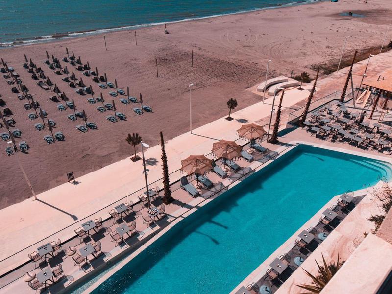 View of a pool and a beach with sun loungers under a blue sky.