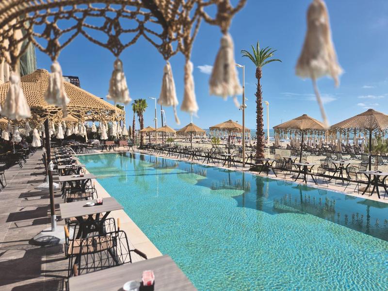 Long outdoor pool with lounge chairs and sun umbrellas under a blue sky at a resort.