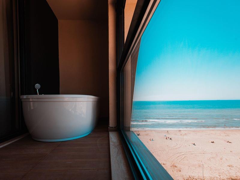 Freestanding bathtub next to a large window overlooking the beach and sea.