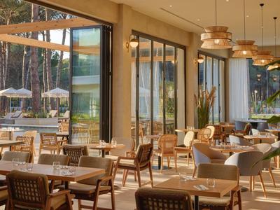 Gemütliches, sonnendurchflutetes Café mit Holz- und Korbmöbeln, großen Fenstern und Blick auf Garten.
